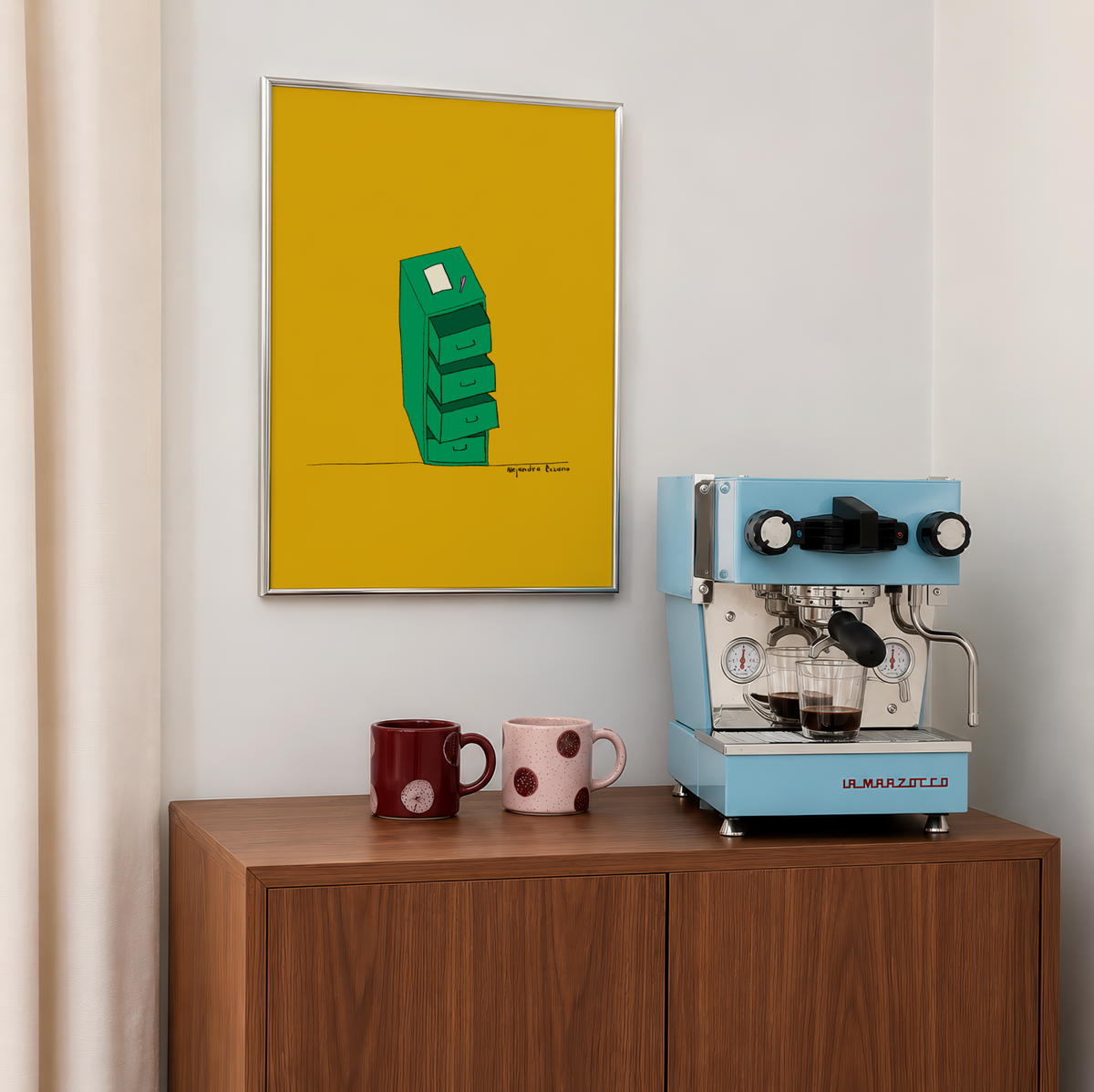 Lámina "Cajón desastre" de Alejandra Lozano con marco plateado colgada sobre un mueble de madera, junto a una cafetera espresso azul claro y dos tazas de lunares granate y rosa.