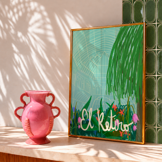 Lámina ilustrada El Retiro con marco dorado junto a un jarrón rosa, con luz y sombras de palmera.