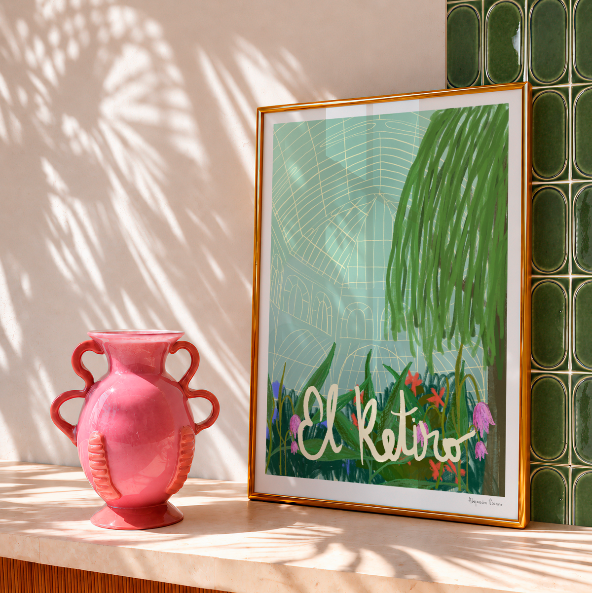 Lámina ilustrada El Retiro con marco dorado junto a un jarrón rosa, con luz y sombras de palmera.