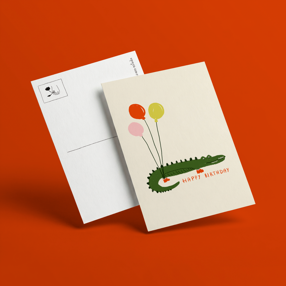 Postal ilustrada de cumpleaños de Alejandra Lozano: cocodrilo verde con botas rojas sujetando tres globos rojo, amarillo y rosa y las palabras Happy Birthday en rojo, junto al reverso blanco de la postal con recuadro para el sello.